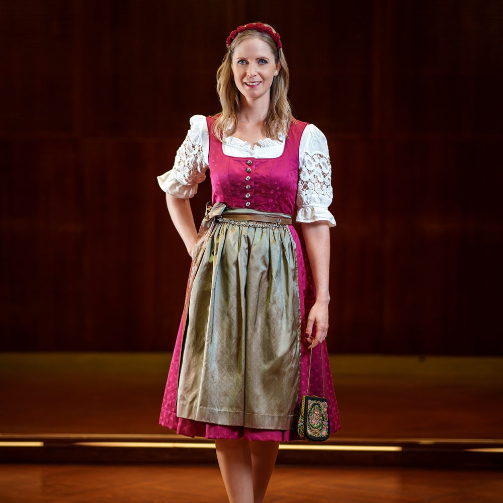 Das Model trägt das Dirndl Mari in der Farbe Berry mit einer reinen Seidenschürze in der Farbe Schilfgrün, passend dazu den Lederblumenhaarreif und eine Retro Trachtentasche